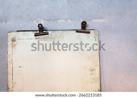 Old worn drawing board with two rusty metal binder clips, mounted against a faded pastel textured wall.
