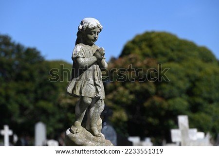 Old, worn, and weathered stone sculpture of a robed child looking down mournfully, hands clasped together in prayer, in a cemetery