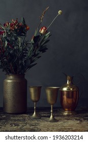 An old worn wooden table topped with a pottery vase with flowers, two bronze goblets and a pitcher. - Shutterstock ID 1936203214