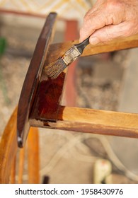 An Old, Wrinkled Hand Holding A Paintbrush Applies Varnish Paint To A Wooden Garden Chair. Painting And Care Of Wooden Furniture. Shallow Depth Of Field