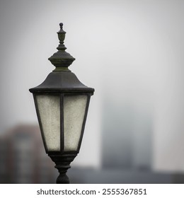 An old wrought iron street lamp stands out in the foreground against a blurred urban background. Its classic and ornate design contrasts with the modernity of the buildings that can be seen. - Powered by Shutterstock