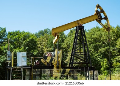 An Old Yellow Oil Rig Pumping Oil Against A Backdrop Of Green Trees And Blue Sky. Oil Extraction From The Well