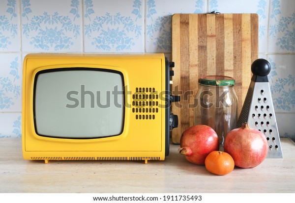 An\
old yellow vintage TV stands in the kitchen next to fruit and\
kitchen utensils against a backdrop of ceramic\
tiles.