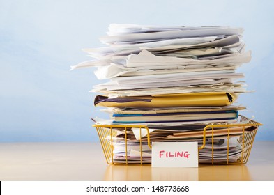 An old yellow wireframe filing tray, piled high with documents and folders, on a light wood veneer desk against light blue background.