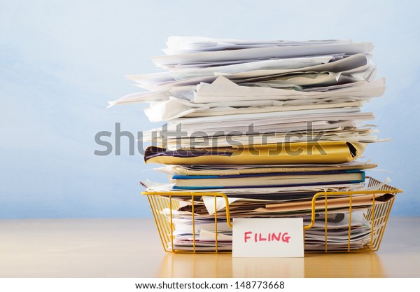 An old yellow wireframe filing tray, piled\
high with documents and folders, on a light wood veneer desk\
against light blue\
background.