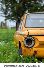 Old Yellow Wrecked Car In Vintage Style. Abandoned Rusty Yellow Car. Close-up Of The Headlights Of The Front View Of A Rusty, Broken, Abandoned Car Near The House. Concept Of Abandoned Used Car.