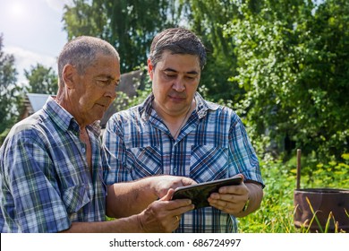 The Old And Young Farmer Are Discussing About The Harvest. They Look At The Tablet And Plan Work On The Farm. The Old Farmer Is Talking With Son.