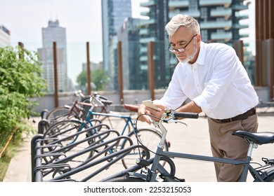 Older 60s man using mobile phone hiring bike in app to ride in the city. Senior old businessman holding smartphone renting bicycle in rental digital mobile application standing in urban park. - Powered by Shutterstock