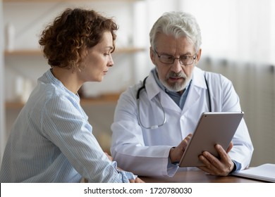 Older adult doctor consulting young female patient using digital tablet tech during checkup appointment meeting. Woman client visiting physician listening diagnosis treatment prescription in hospital. - Powered by Shutterstock