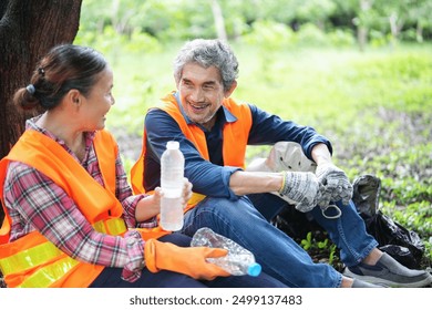 older adult husband and wife in safety vest resting under a tree and drinking water,asian senior couple spend holiday time doing volunteer activity collecting and sorting waste in the park - Powered by Shutterstock