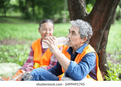 older adult husband and wife in safety vest resting under a tree and drinking water,asian senior couple spend holiday time doing volunteer activity collecting and sorting waste in the park - Powered by Shutterstock