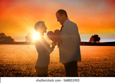 Older Affectionate Couple Holding Pink Heart Shape Against Countryside Scene