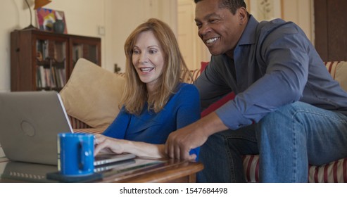 Older African American And Caucasian Husband And Wife Using Laptop Computer Together In Living Room. Mature Homeowner Married Couple Smiling While Planning Trip Online