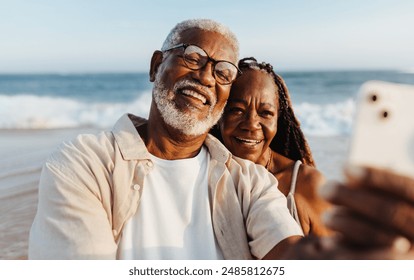 An older African American couple shares a joyful moment, taking a selfie together with the ocean backdrop during a serene beach walk. - Powered by Shutterstock