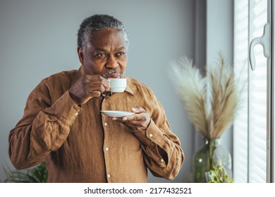 Older African Man Drinking Coffee While Standing By The Window In The Morning, Happy Older African Man Holding A Cup Of Coffee At Home, Retired Old People Healthy Lifestyle. Retirement Lifestyle