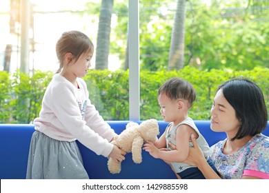 Older Asian sister give teddy bear toy to younger brother with mother take care her children. Family relationship concept.