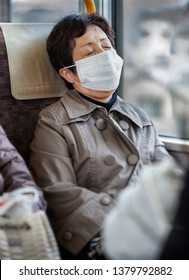 Older Asian Woman Wearing A Tan Raincoat And Surgical Pollution Mask Sitting On A Bus Train Subway