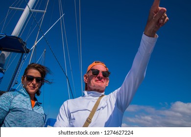 Older Baby Boomer Caucasian Couple With Man Pointing Into The Distance. Deep Blue Sky With Sailboat Rigging In Background. Woman's Hair Blowing In The Wind. Both Wearing Sunglasses