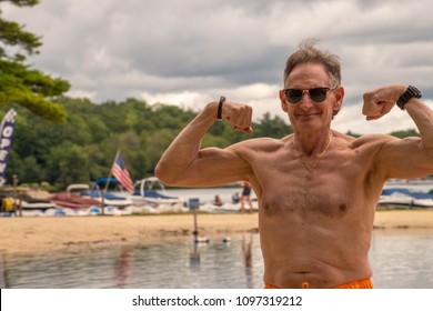 Older Baby Boomer Wearing Sunglasses Topless In Bathing Suit Flexing His Muscles With Lakes And Boats In Background