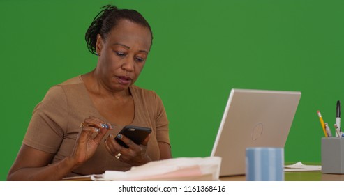 Older Black Woman Uses Her Phone While Sitting At Her Work Desk On Green Screen