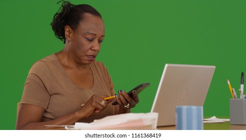 An Older Black Woman Works Using Phone At Her Workstation On Green Screen