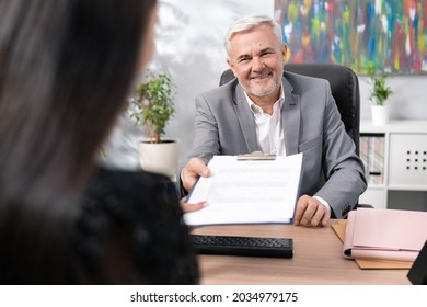 Older Boss In Suit Sits In Office Chair At Desk, Conducts Job Interview, Man Is Pleased With Woman, Hands Her Employment Contract To Sign, Smiles Both Hold Up Pad Of Document