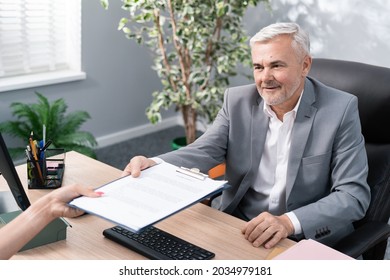 Older Boss In Suit Sits In Office Chair At Desk, Conducts Job Interview, Man Is Pleased With Woman, Hands Her Employment Contract To Sign, Smiles Both Hold Up Pad Of Document