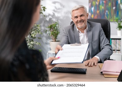 Older Boss In Suit Sits In Office Chair At Desk, Conducts Job Interview, Man Is Pleased With Woman, Hands Her Employment Contract To Sign, Smiles Both Hold Up Pad Of Document