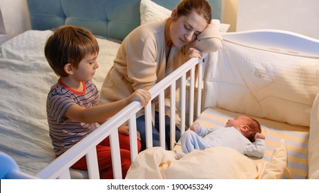 Older Boy Helping His Tired Mother Rocking Baby Ccrib In Bedroom At Night. Children Helping Parents. Motherhood And Sleepless Nights.