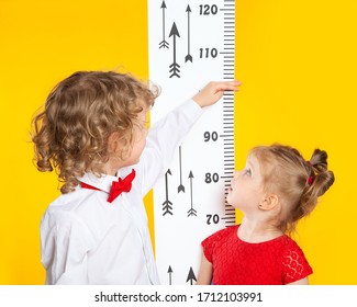 The Older Brother Measures The Height Of The Younger Sister Using A Height Meter. Slavic, European Children On A Yellow Background. A Boy In A Red Bow Tie. A Girl In A Red Dress.