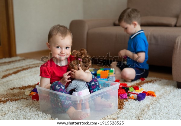 The older brother is playing on the carpet\
with the constructor, and the sister is sitting in a box from under\
the constructor.