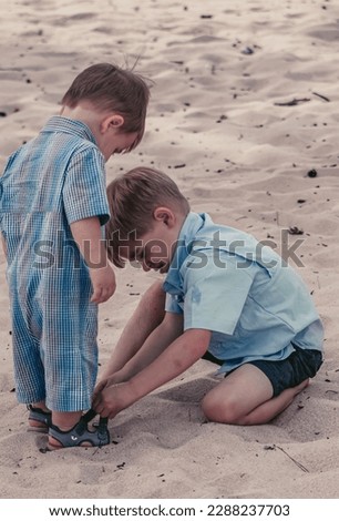 older brother sitting down helping the younger with his sandals while walking on the beach. showing love and caring relationship between siblings