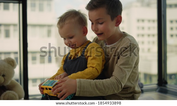 Older brother\
teaching younger play with toy car in in living room. Little kid\
sitting in elder boy laps on floor at home interior. Children\
playing toy monster truck\
indoors.