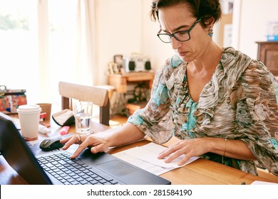 Older Business Woman Busy Entering Digits On Her New Laptop Computer From A Printed List That She's Reading While Wearing Glasses