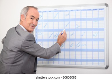 Older Businessman Writing On A Wall Planner