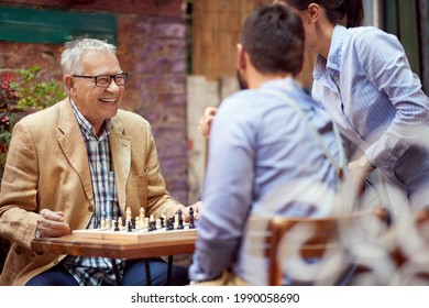 Older Caucasian Male Smiling, Happy To Win In Chess Game. Sitting In Outdoor Cafe With His Younger Opponent Who Is Talking To Female.