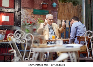 Older Caucasian Male Smiling With Thumbs Up. Young Guy With His Hand Over His Eyes. Sitting In Outdoor Cafe, Playing Chess. Winning And Loosing Concept