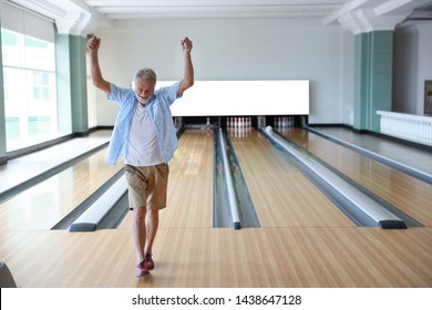 Older Caucasian Men, White Beard And White Hair In White And Blue Shirt Playing Bowling And He Put His Hands Up After Finish The Game Meaning Of The Winner In Sport Club