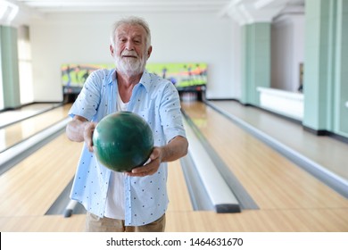 Older Caucasian Men, White Beard And White Hair In White And Blue Shirt Playing Bowling While He Holding Bowling Ball To Someone In Sport Club