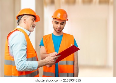 Older Construction Supervisor With Younger Worker Helper At Construction Site. Managers Wearing Protective Workwear, Hard Hat, Construction Scaffolding. Construction Workforce, Working Labor Man.