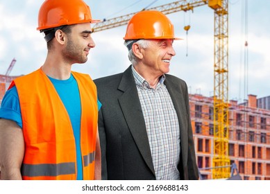 Older Construction Supervisor With Younger Worker Helper At Construction Site. Managers Wearing Protective Workwear, Hard Hat, Construction Cranes On Skyline. Construction Workforce, Working Labor Man