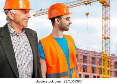 Older Construction Supervisor With Younger Worker Helper At Construction Site. Managers Wearing Protective Workwear, Hard Hat, Construction Cranes On Skyline. Construction Workforce, Working Labor Man