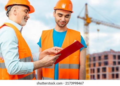 Older Construction Supervisor With Younger Worker Helper At Construction Site. Managers Wearing Protective Workwear, Hard Hat, Construction Cranes On Skyline. Construction Workforce, Working Labor Man