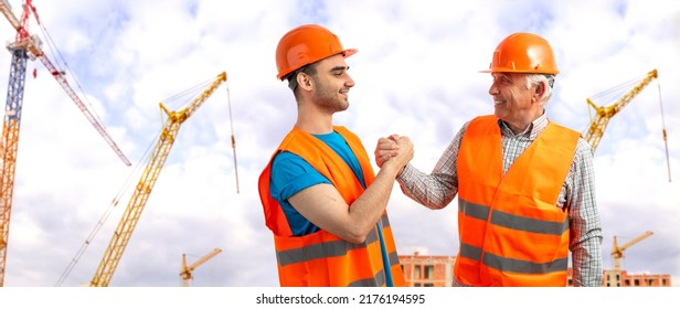 Older Construction Supervisor With Younger Worker Helper At Construction Site. Managers Wearing Protective Workwear, Hard Hat, Construction Cranes On Skyline. Construction Workforce, Working Labor Man