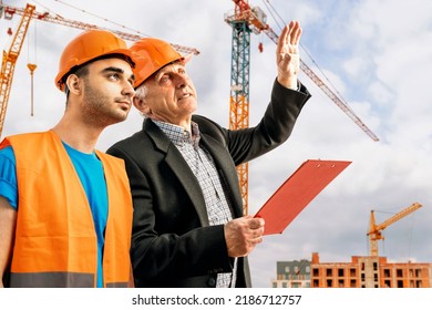 Older Construction Supervisor With Younger Worker Helper Construction Site. Managers Wearing Protective Workwear, Hard Hat, Construction Cranes On Skyline. Construction Workforce, Working Labor Man.