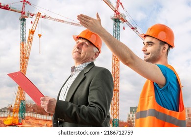 Older Construction Supervisor With Younger Worker Helper Construction Site. Managers Wearing Protective Workwear, Hard Hat, Construction Cranes On Skyline. Construction Workforce, Working Labor Man.