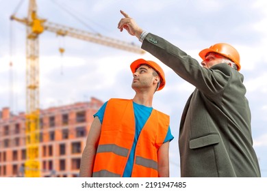 Older Construction Supervisor With Younger Worker Helper At Construction Site. Managers Wearing Protective Workwear, Hard Hat, Construction Cranes On Skyline. Construction Workforce Working Labor Man.