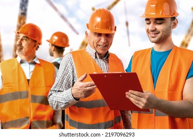 Older Construction Supervisor With Younger Worker Helper At Construction Site. Managers Wearing Protective Workwear Hard Hat, Construction Cranes On Skyline. Construction Workforce, Working Labor Man.