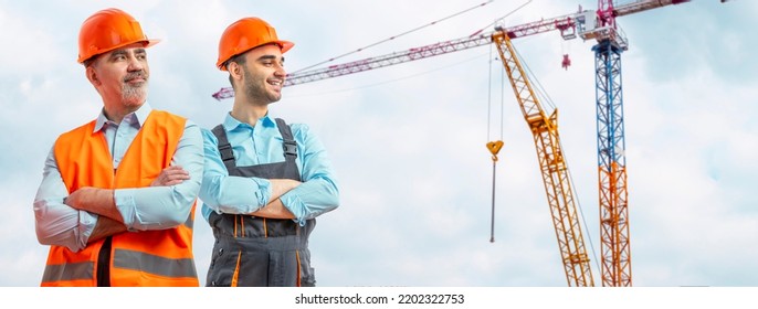 Older Construction Supervisor With Younger Worker Helper At Construction Site. Managers Wearing Protective Workwear, Hard Hat, Construction Cranes Skyline. Construction Workforce, Working Labor Man.