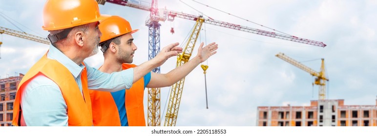 Older Construction Supervisor With Younger Worker Helper Construction Site. Managers Wearing Protective Workwear, Hard Hat, Construction Cranes On Skyline. Construction Workforce, Working Labor Man.